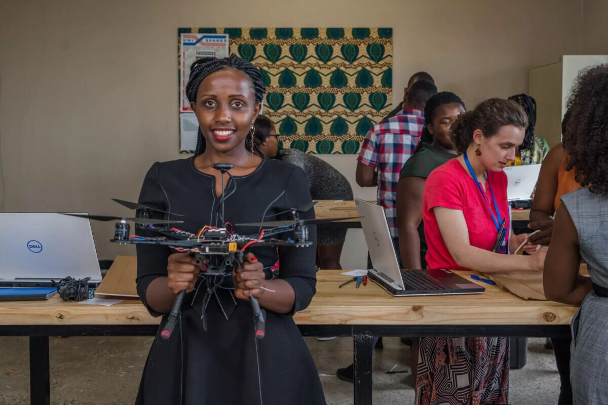 A woman holds a multi-rotor drone while participants work with laptops and equipment at a technology workshop.