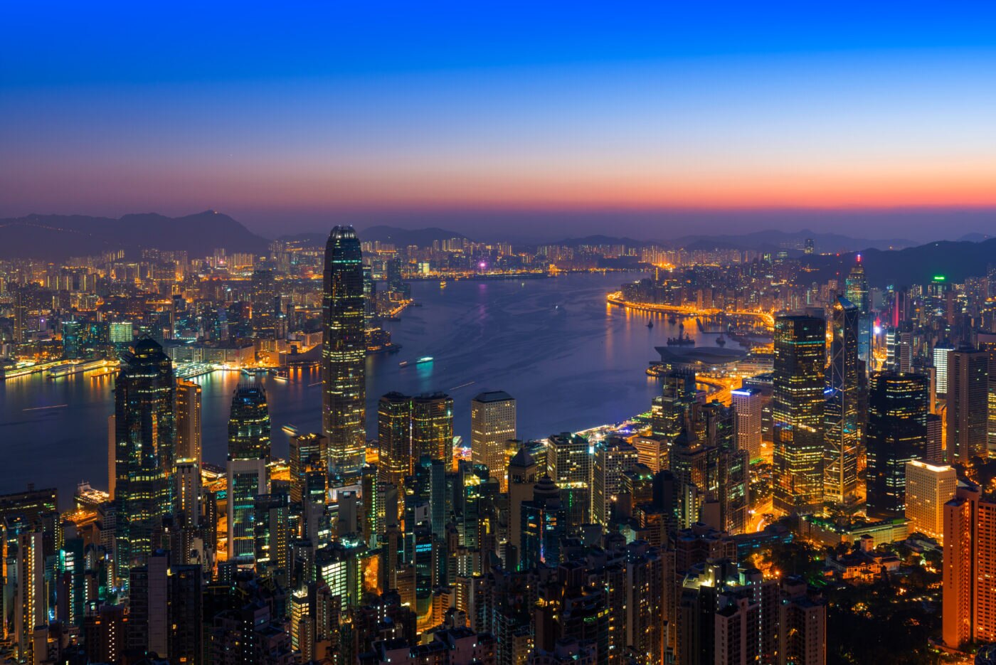 Hong Kong skyline and Victoria Harbour at dusk, with illuminated high-rise buildings and the city stretching across the waterfront.